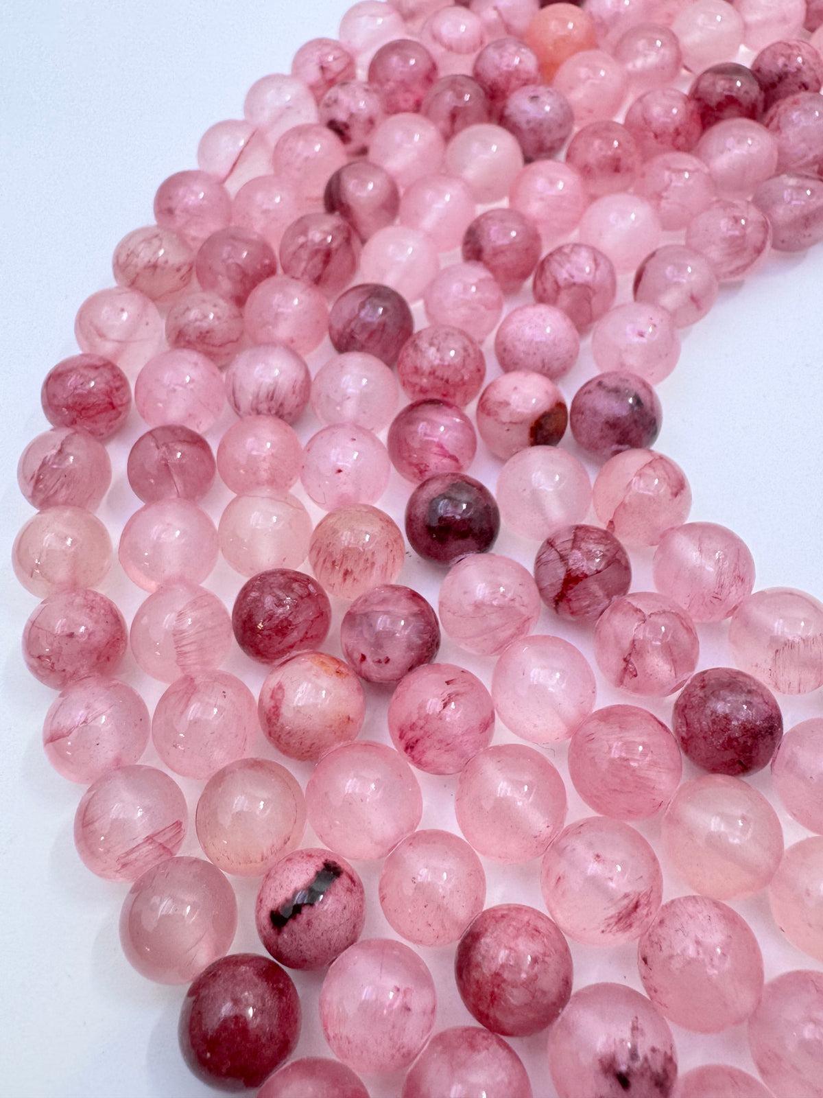 Multiple strands of polished round pink persian jade gemstone beads, showing natural pink and reddish tones with some darker inclusions, arranged on a plain white background.
