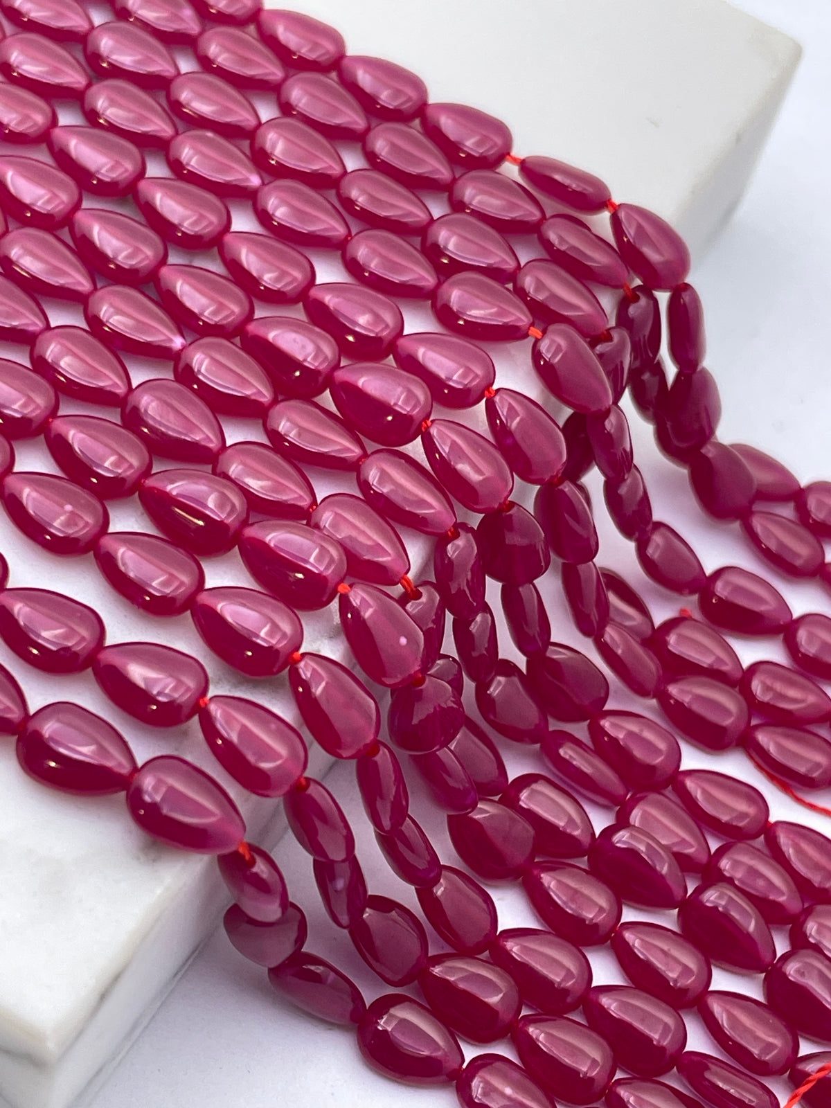 A close-up of glossy, teardrop-shaped lab ruby beads in a deep magenta-pink color, draped over a white marble block on a white surface.