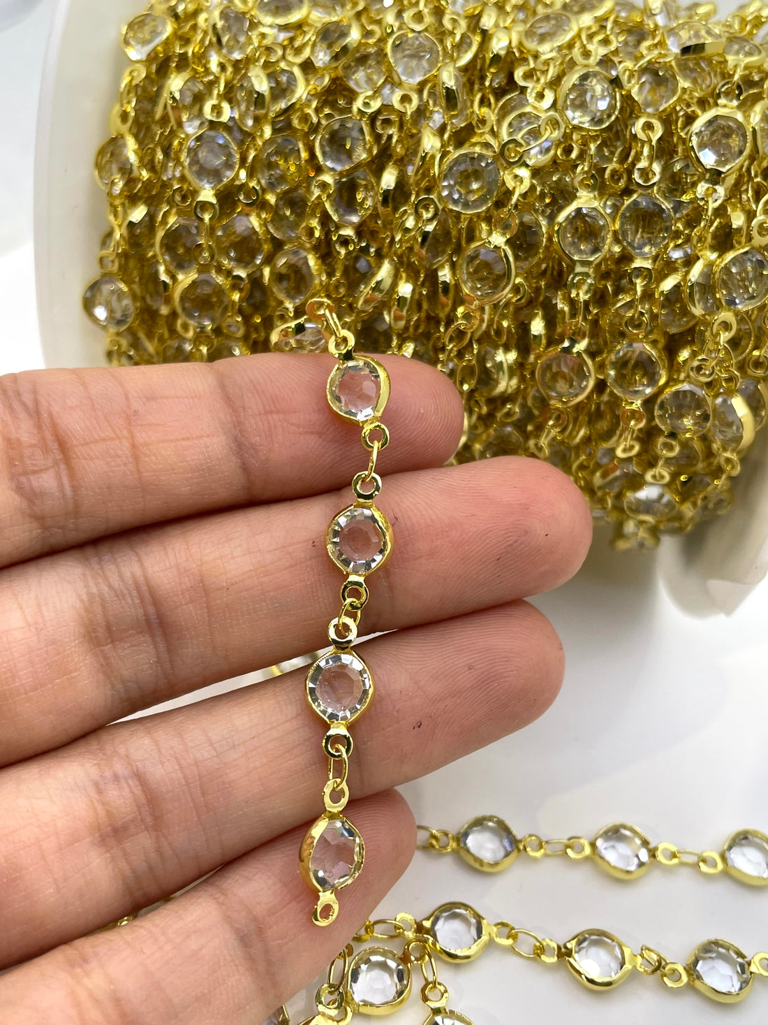 Gold chain trim embellished with clear faceted rhinestones, shown with a spool in the background and several draped strands in the foreground on a white surface.