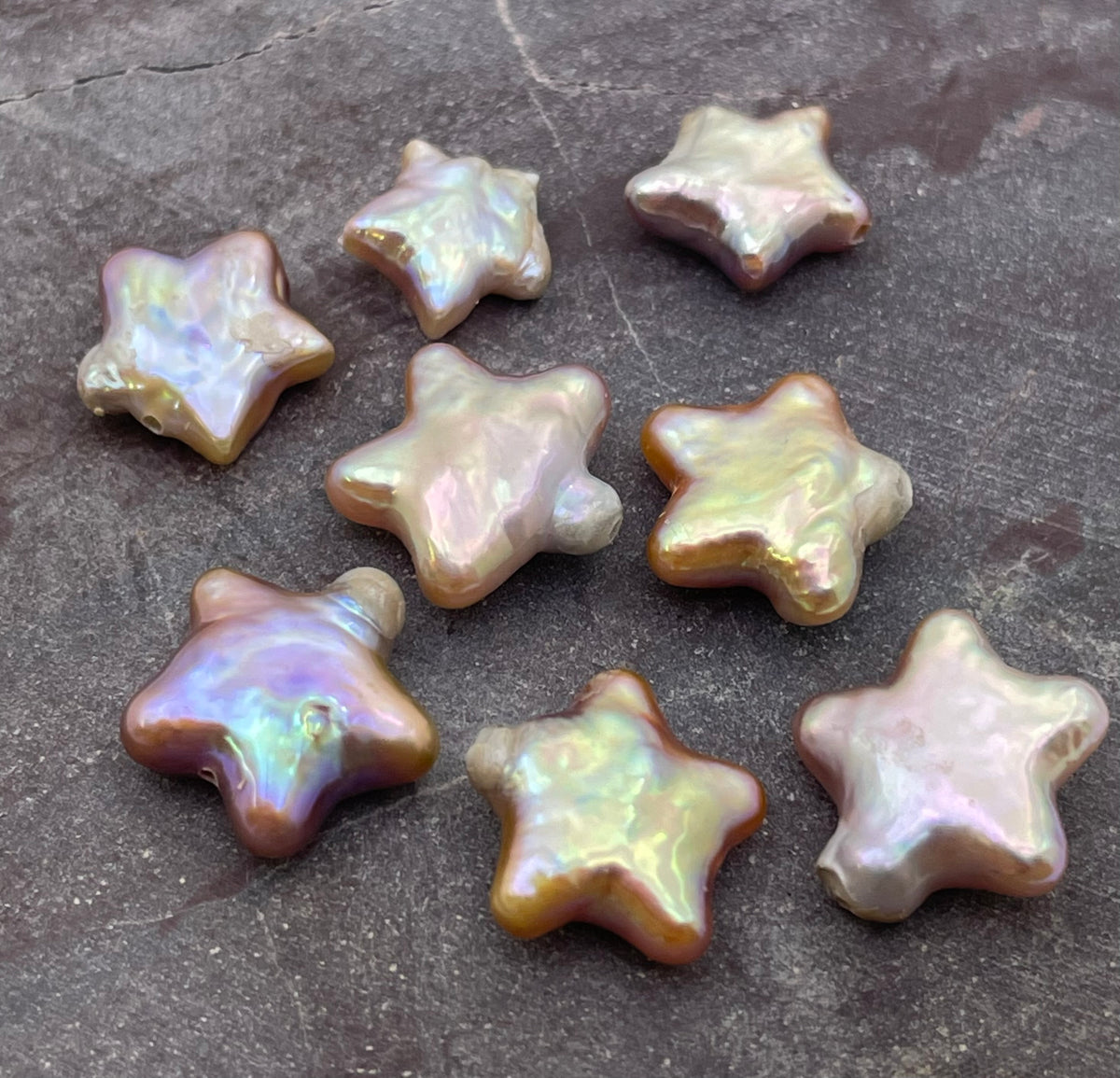 Seven star-shaped baroque pearls with iridescent, pearlescent finishes in shades of beige, gold, and soft purple, arranged on a dark textured stone surface.