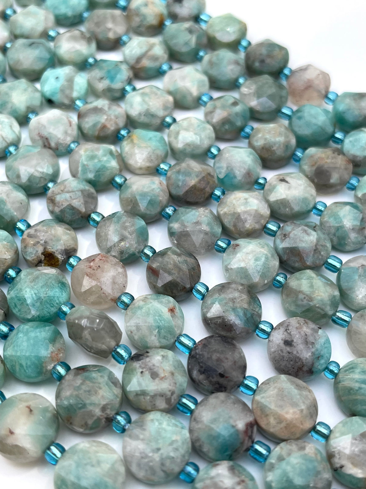 Faceted round amazonite beads in a mottled light blue-green, gray, and beige stone pattern, strung on a cord with small metallic blue spacer beads, shown on a white background.