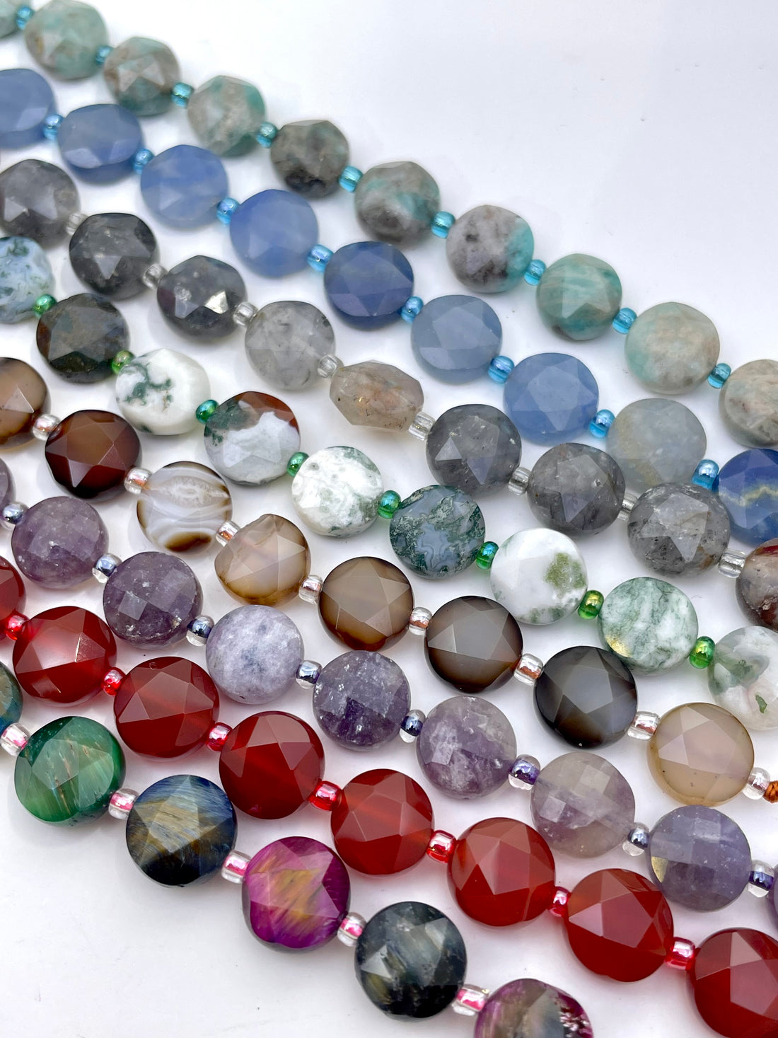 A close-up of multiple strands of faceted gemstone beads held in a hand. The beads come in many colors and patterns including red, purple, blue, gray, green, white, and banded agate designs.