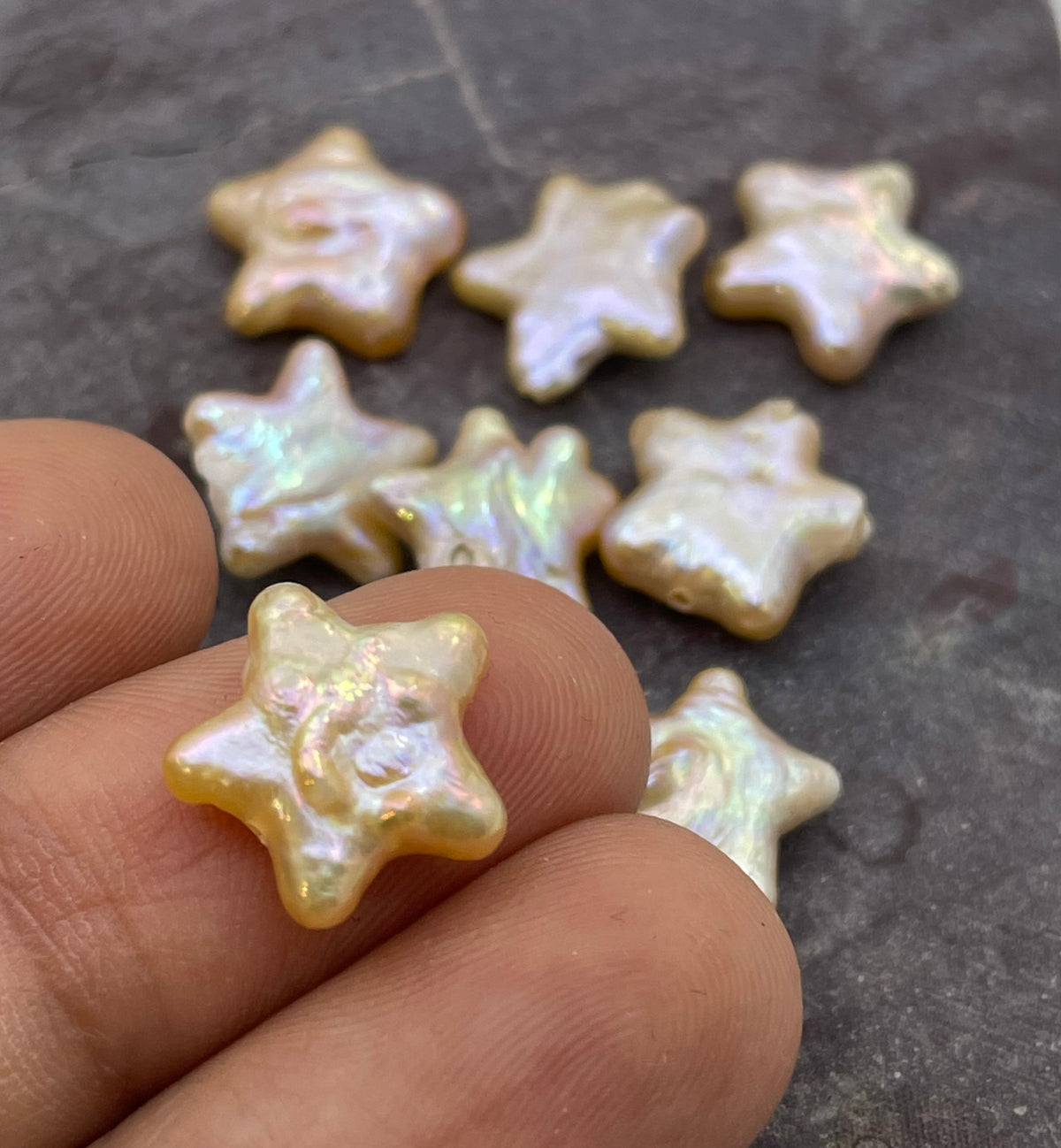 Nine small star-shaped beads with an iridescent pearl-like finish are displayed on a dark textured surface, with several held between fingers to show scale.