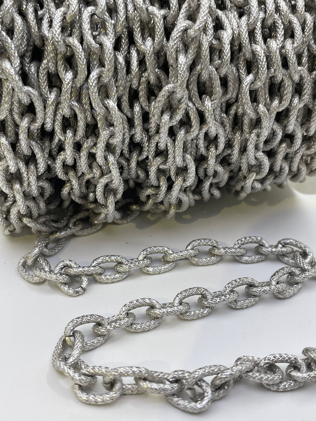 A close-up of a silver metallic chain with textured, rope-like links, shown coiled in the background and draped in the foreground on a white surface.