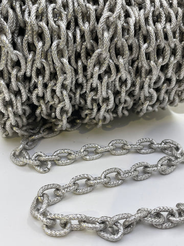 A close-up of a silver metallic chain with textured, rope-like links, shown coiled in the background and draped in the foreground on a white surface.