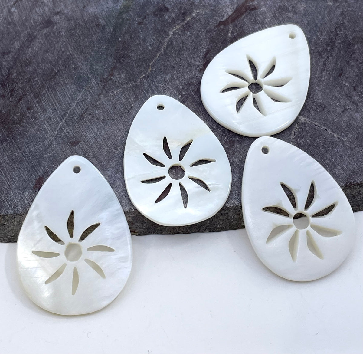 Four white teardrop-shaped shell pendants with starburst cutouts, arranged on a split white and dark gray background.