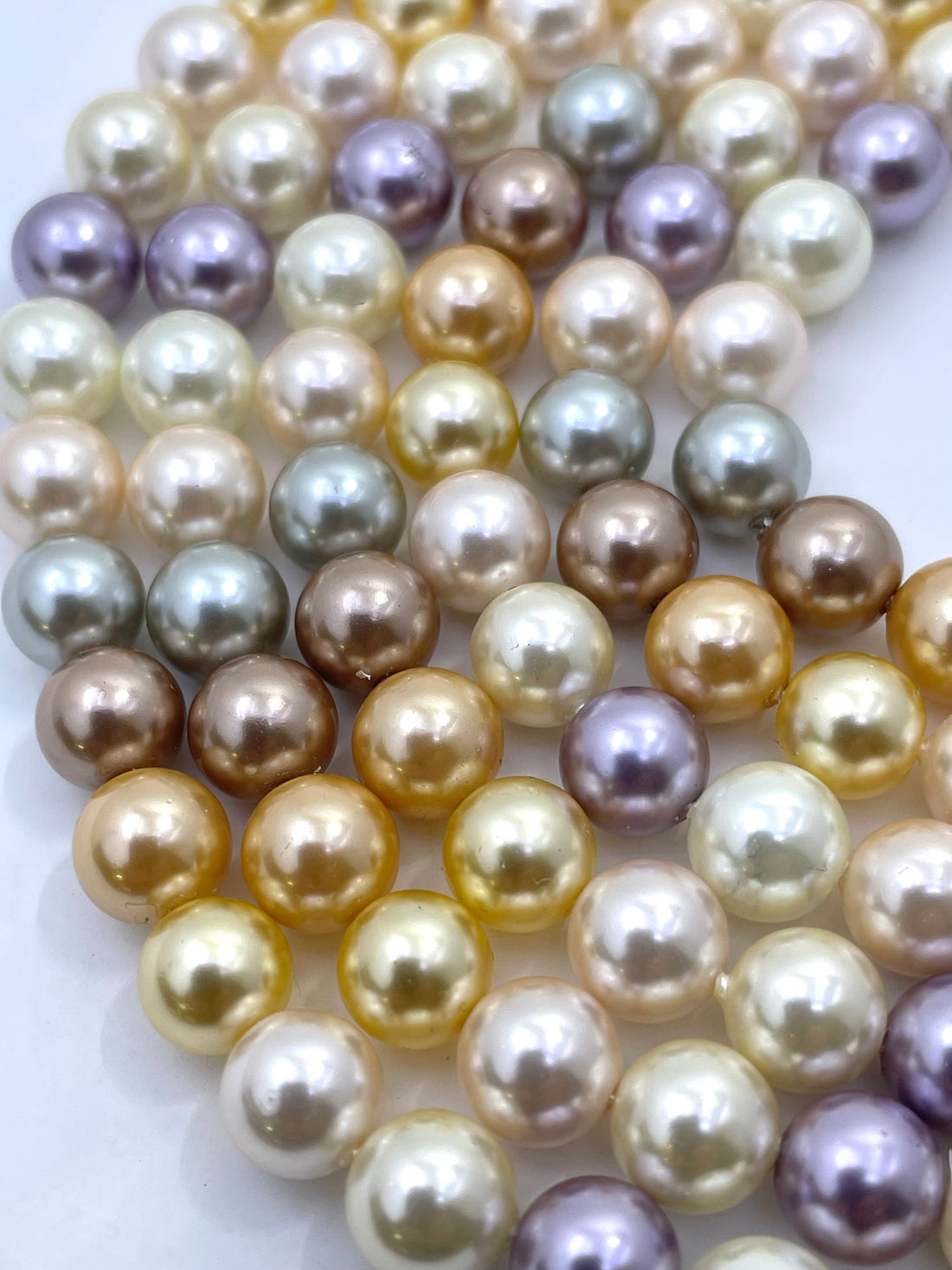 Close-up of strands of shell pearl beads in a mix of champagne gold, cream, white, taupe, and lavender purple on a white background.