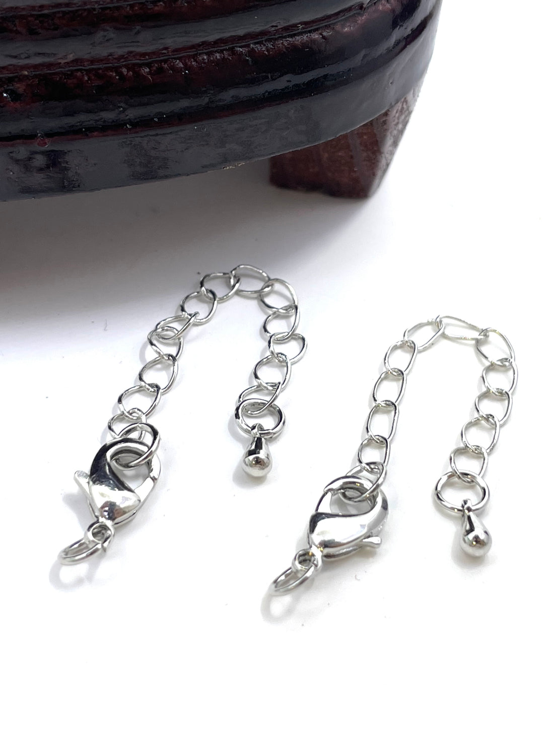 Two silver metal chain extender clamps with lobster clasps and small ball charms, displayed on a white surface with a dark wooden object partially visible at the top.
