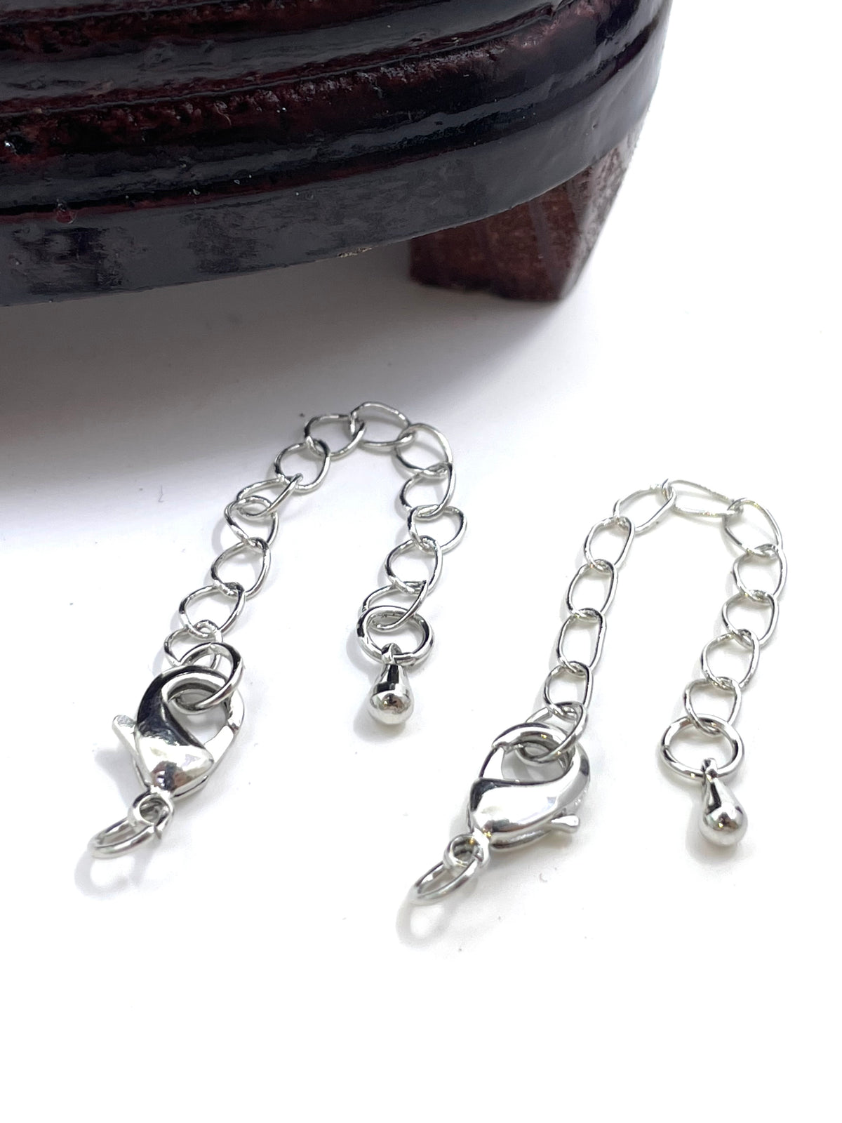 Two silver metal chain extender clamps with lobster clasps and small ball charms, displayed on a white surface with a dark wooden object partially visible at the top.