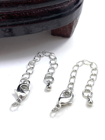 Two silver metal chain extender clamps with lobster clasps and small ball charms, displayed on a white surface with a dark wooden object partially visible at the top.