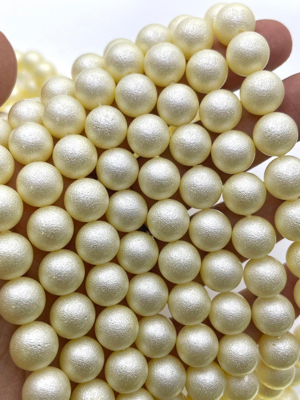 A close-up of many small round shell pearl beads in a warm ivory or cream tone, held in a hand against a plain light background.
