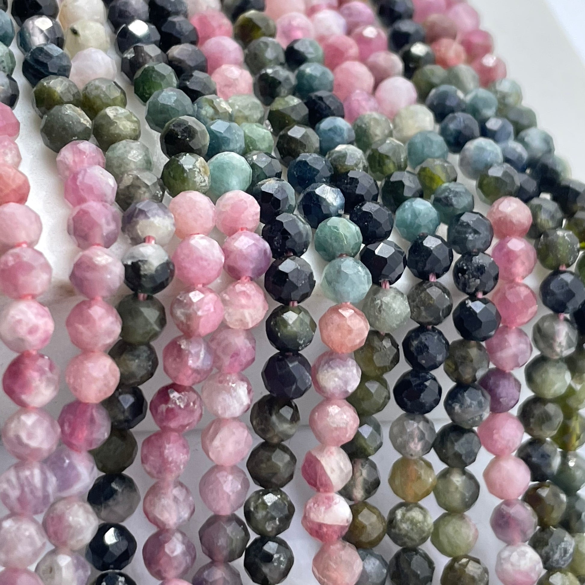 Multiple strands of faceted round rainbow tourmaline gemstone beads in assorted colors, including pink, green, blue, gray, and black, arranged closely together on a white background.