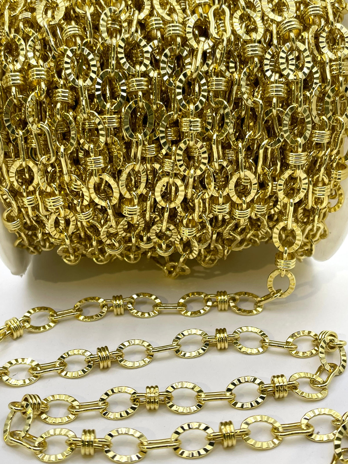 A close-up of a gold-colored chain link accessory, showing a spool of the chain in the background and several individual links draped in the foreground on a white surface.