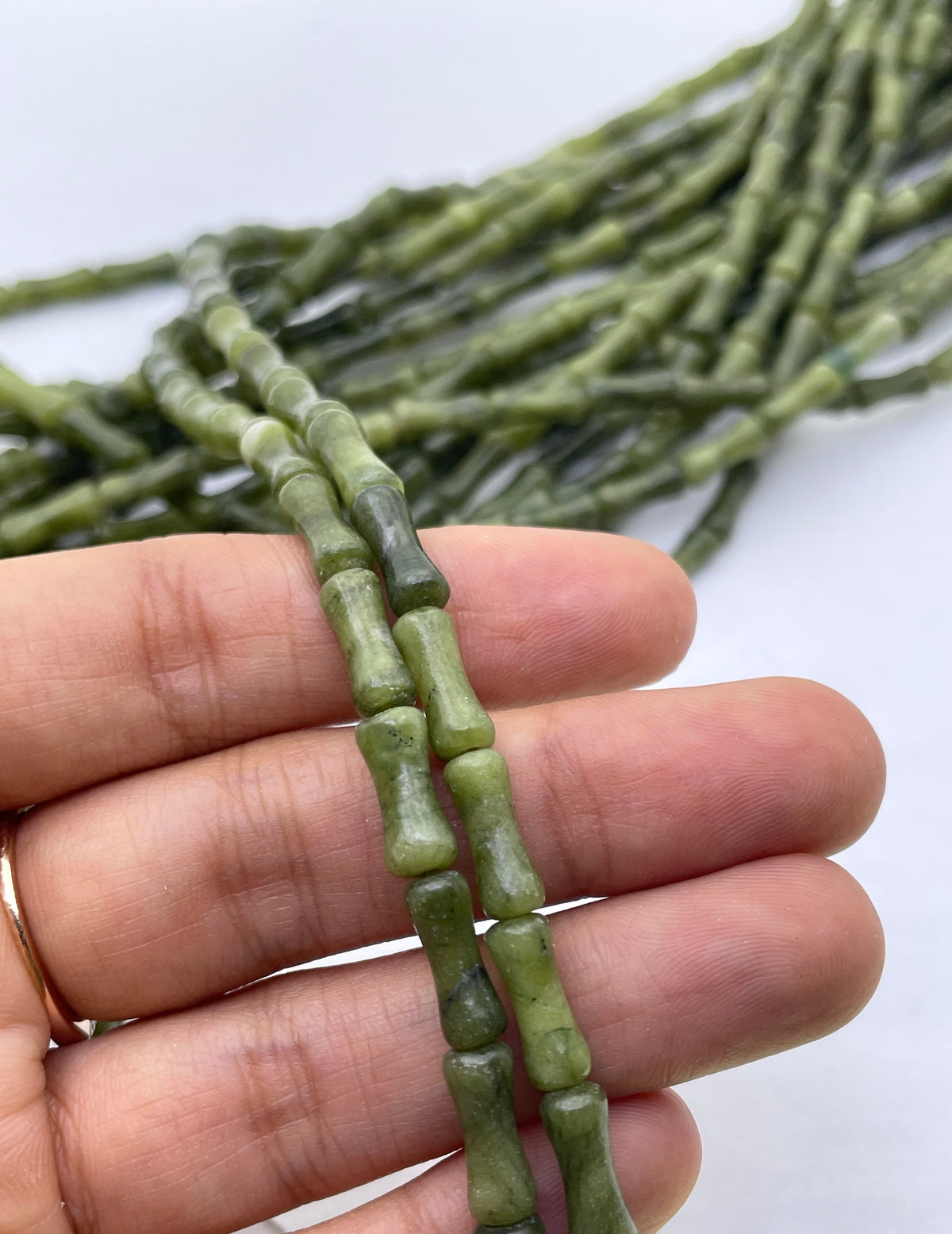 A hand holding two strands of green bamboo-style nephrite jade beads, with more of the same strands blurred in the background on a white surface.
