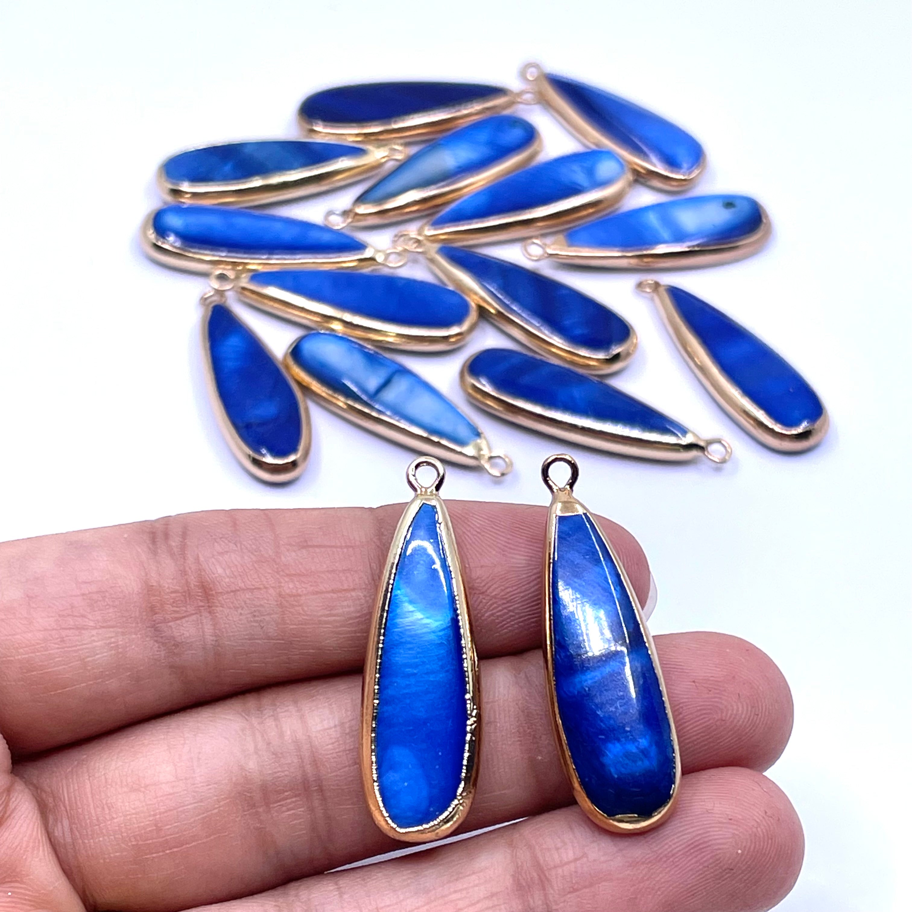 Blue teardrop-shaped pendant charms with gold bezel settings, shown with two held in a hand and a group of nine more in the background on a white surface.