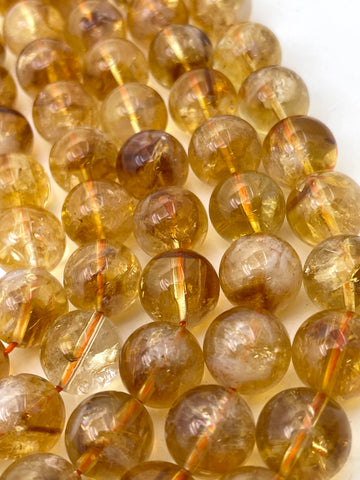 Close-up of round translucent amber-yellow citrine beads strung on cords, showing internal inclusions and a glossy finish against a white background.