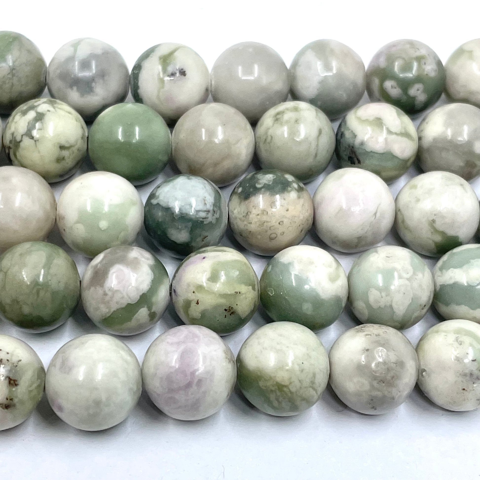 A close-up of polished round peace jade gemstone beads or beads arranged in rows on a white background. The beads are predominantly light gray and white with natural green and beige marbling.