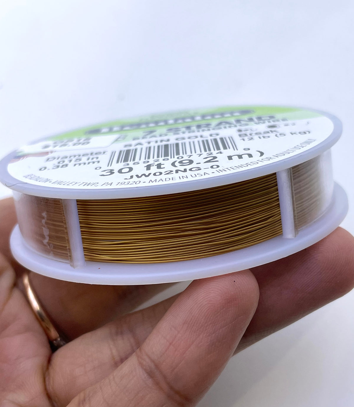 A hand holding a spool of JWG2NA Gold Tinsel wire, showing the golden-colored wire wound around the plastic spool against a plain white background.