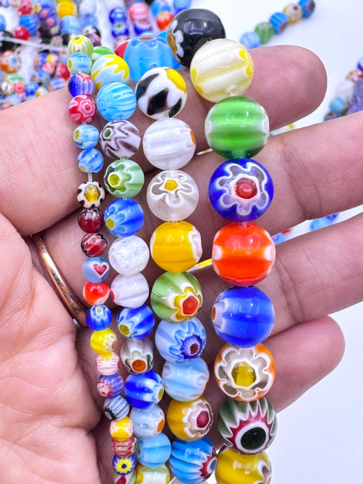 A hand holding several strands of colorful glass bead strands, featuring various patterns and colors like blue, green, yellow, orange, white, and black.