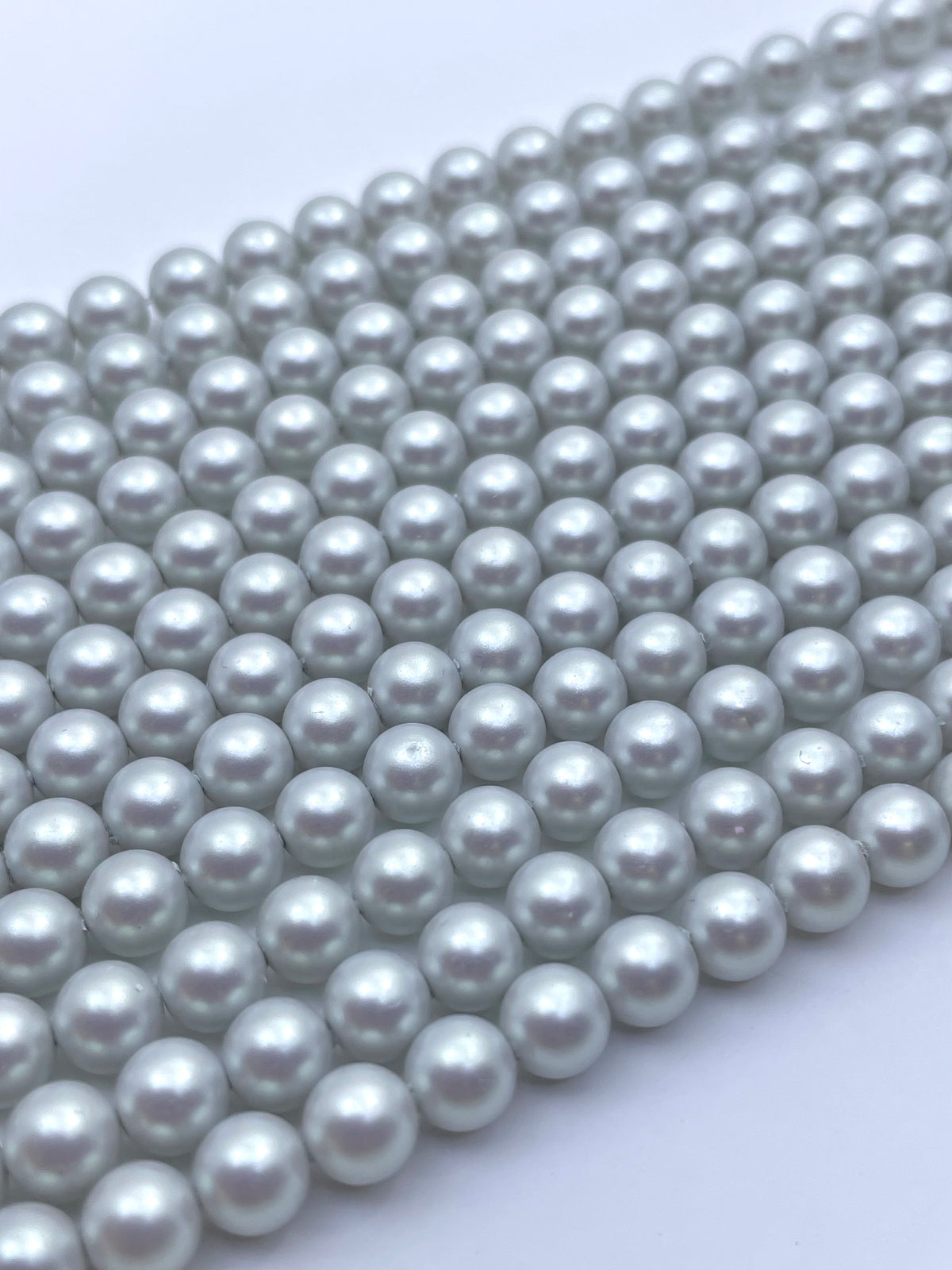 A close-up of multiple strands of small, round, lustrous shell pearl beads in a light silver-gray color, arranged diagonally on a plain white background.