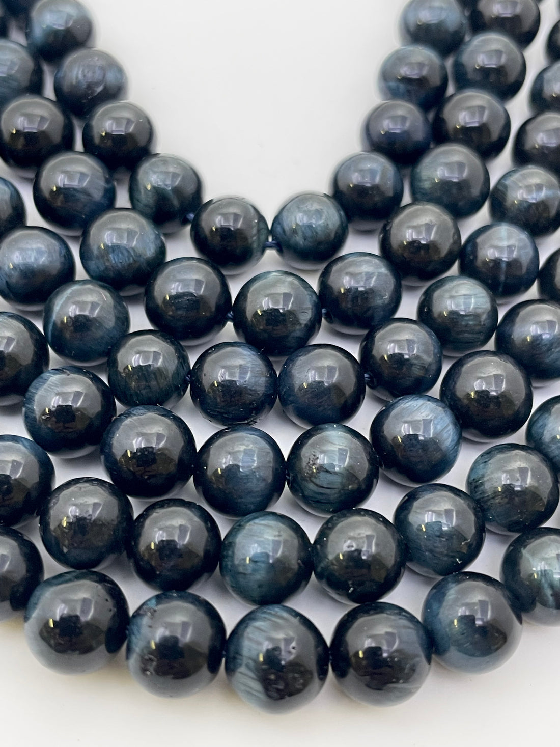 A close-up of glossy dark blue to black round tiger eye beads arranged in strands on a plain white background.