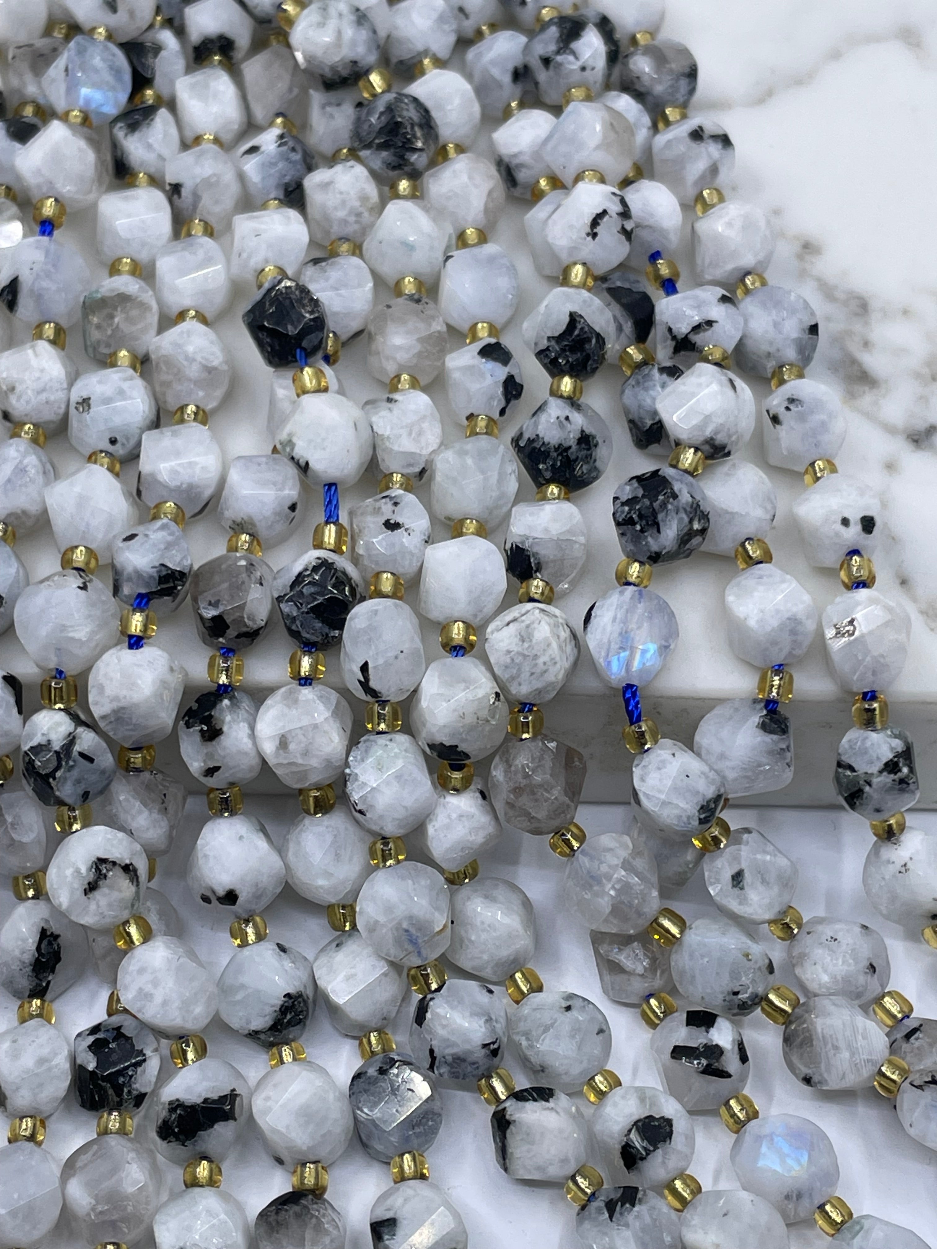A close-up of multiple strands of faceted rainbow moonstone with black inclusions and small gold spacer beads, laid out on a white marble surface.