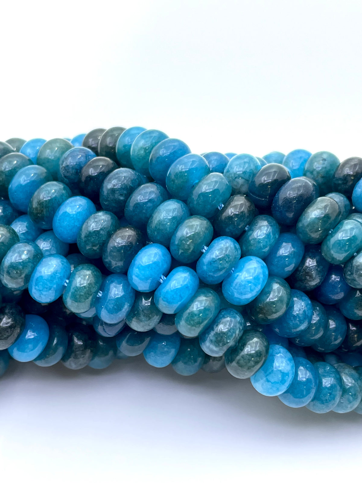 A close-up of multiple strands of smooth donut apatite jade beads in shades of bright blue, teal, and dark green, arranged on a plain white background.