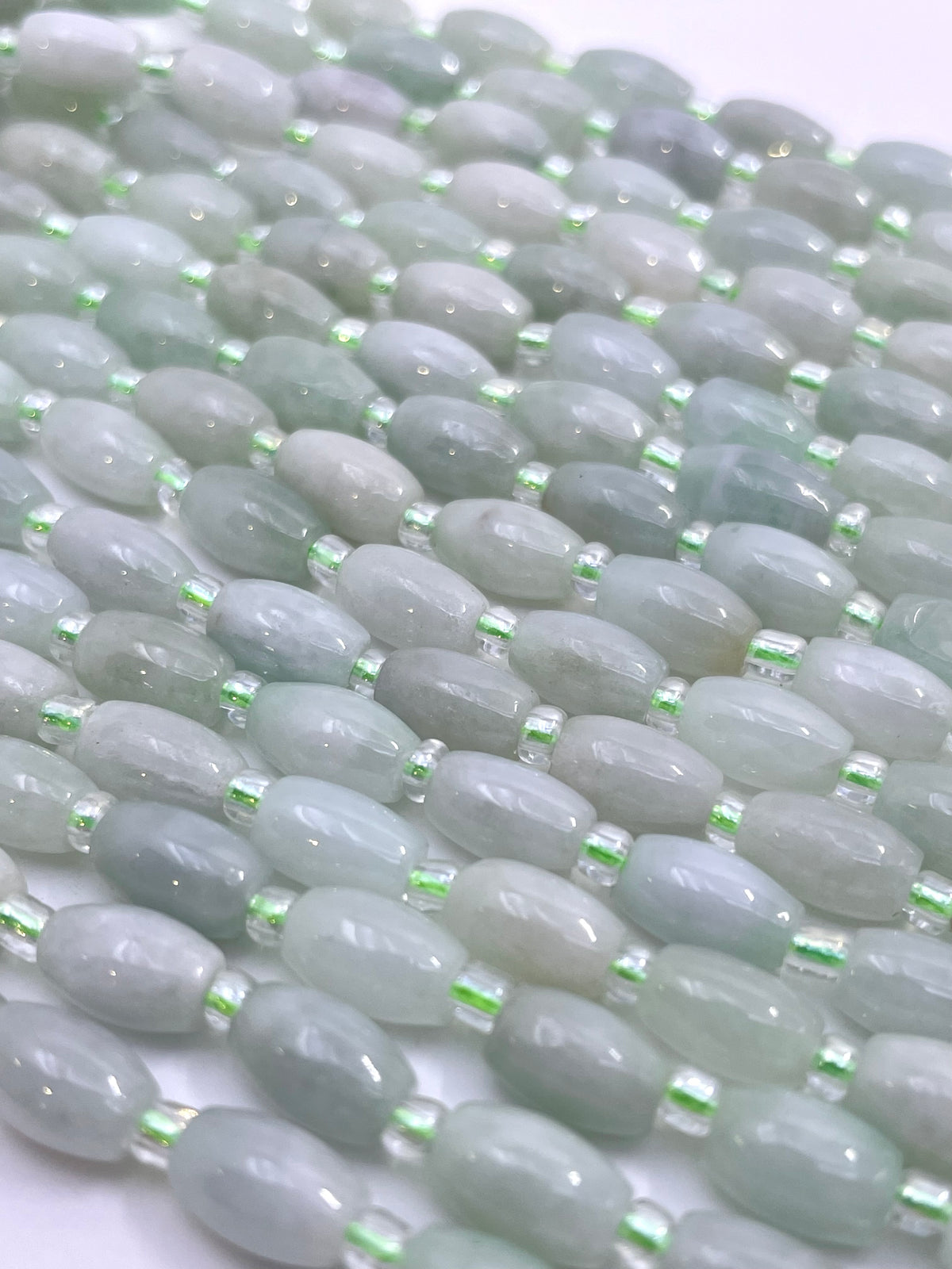 Close-up of multiple strands of burma jade oval beads in pale green, mint, and soft gray tones, separated by small metallic silver spacer beads, on a white background.