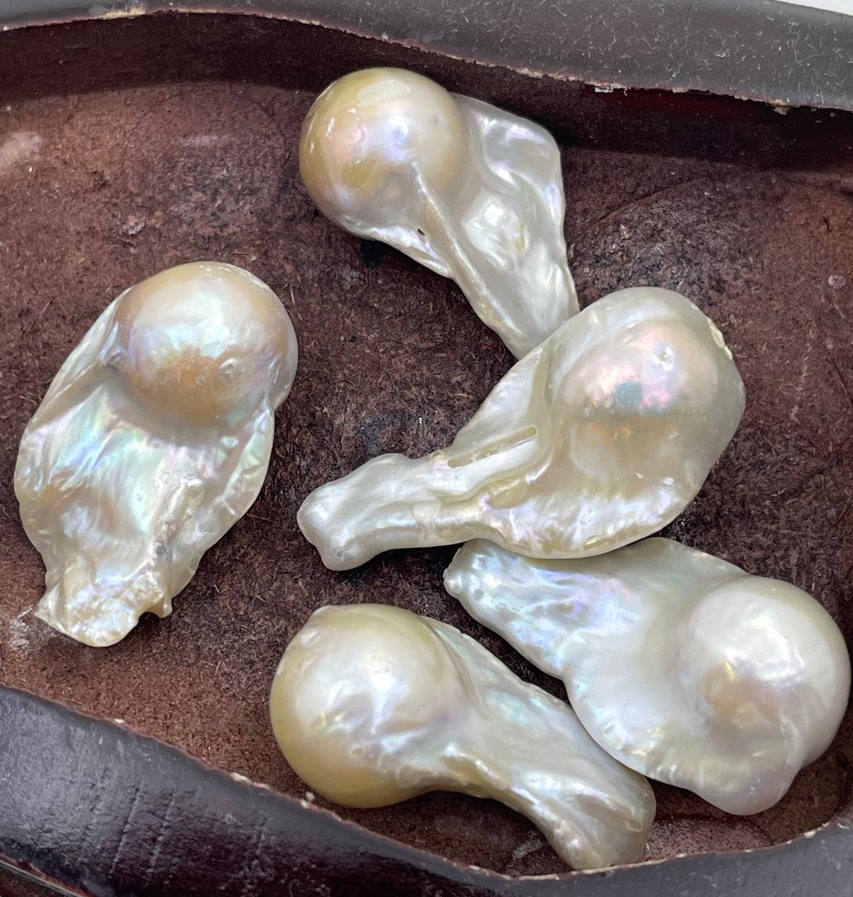 Five irregular, bumpy freshwater pearl pieces with a pearly off-white iridescent sheen resting on a dark brown textured surface.