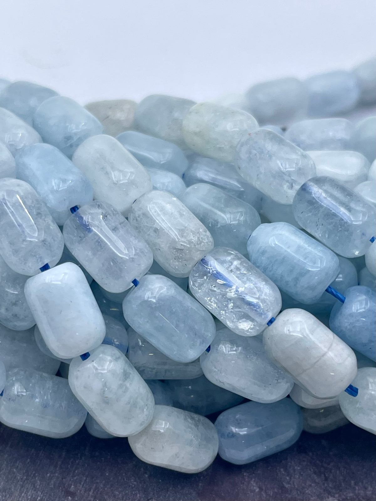 A pile of light blue and white aquamarine gemstone beads with a rounded rectangular shape, strung on a dark cord.