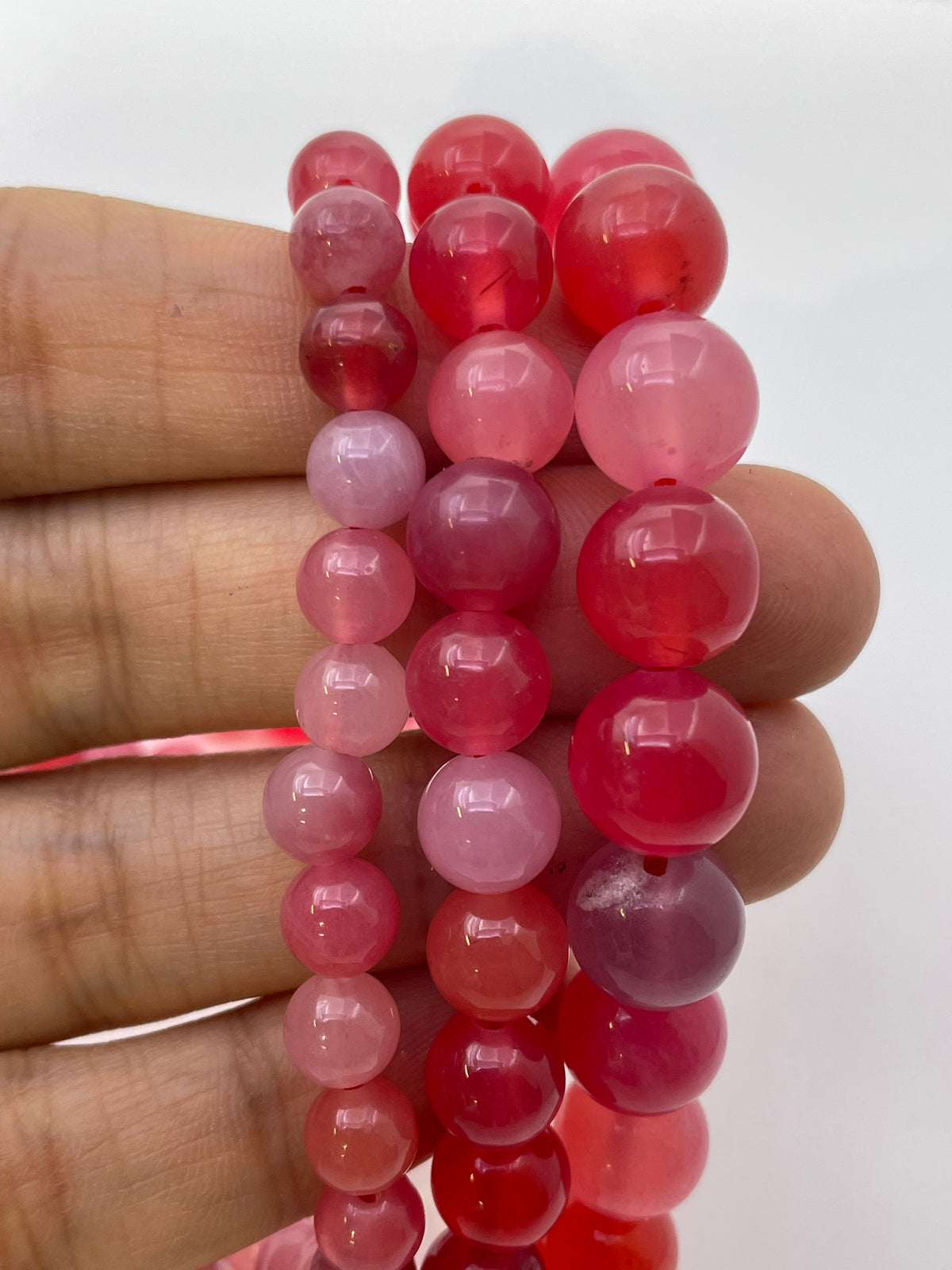 A close-up of a hand holding three sizes  of translucent round scarlet agate beads in shades of pink and red, against a plain white background.