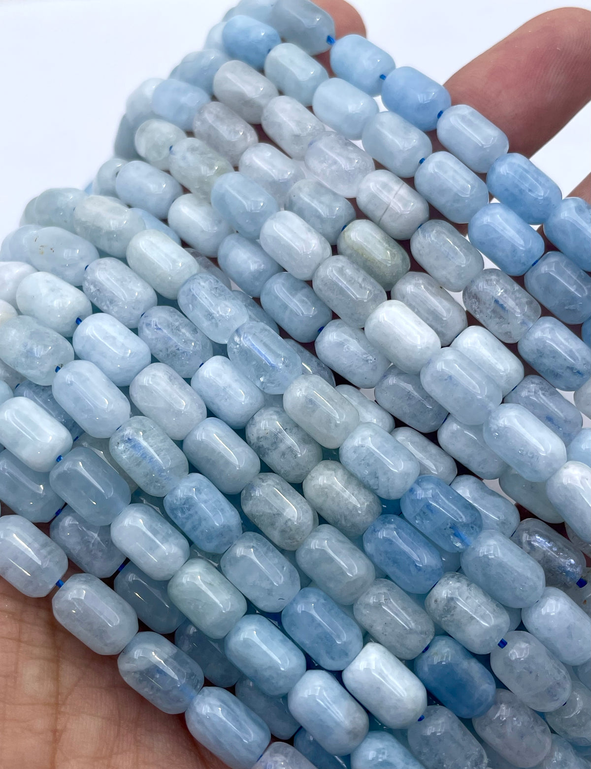 Close-up of multiple strands of light blue and white aquamarine cylindrical beads held in a hand against a plain white background.