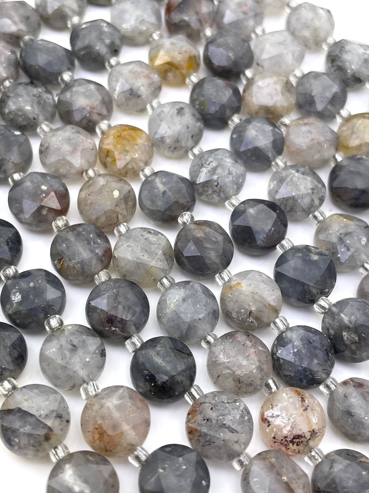 Faceted round grey rutilated quartz gemstone beads in gray, charcoal, black, and smoky tones with small silver spacer beads, arranged in rows on a white background.