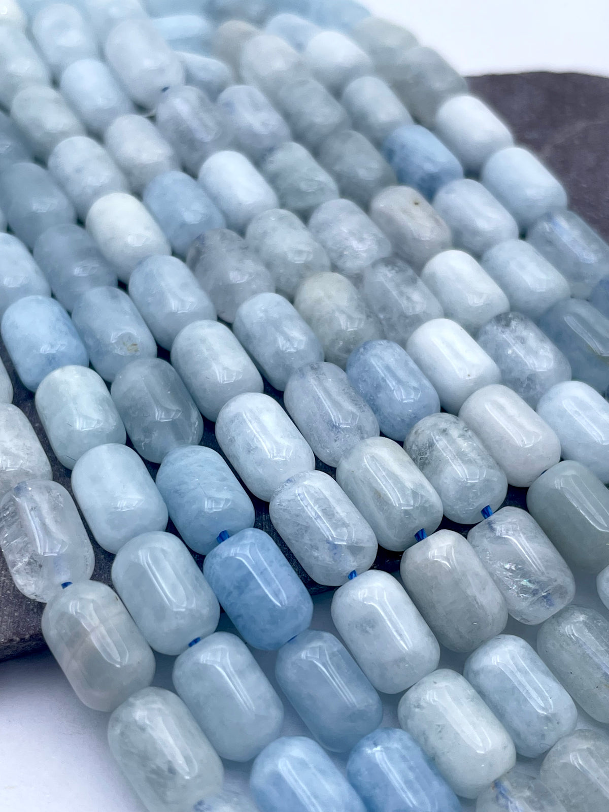 Close-up of a strand of polished light blue and white cylindrical aquamarine beads, arranged diagonally on a neutral surface.