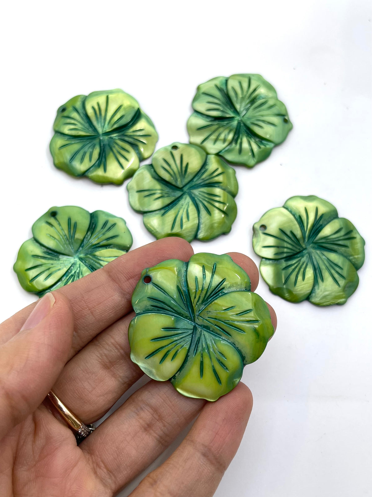Five green leaf-shaped mother of pearl flower pendant with dark green veining, displayed on a white background with one being held in a hand for scale.