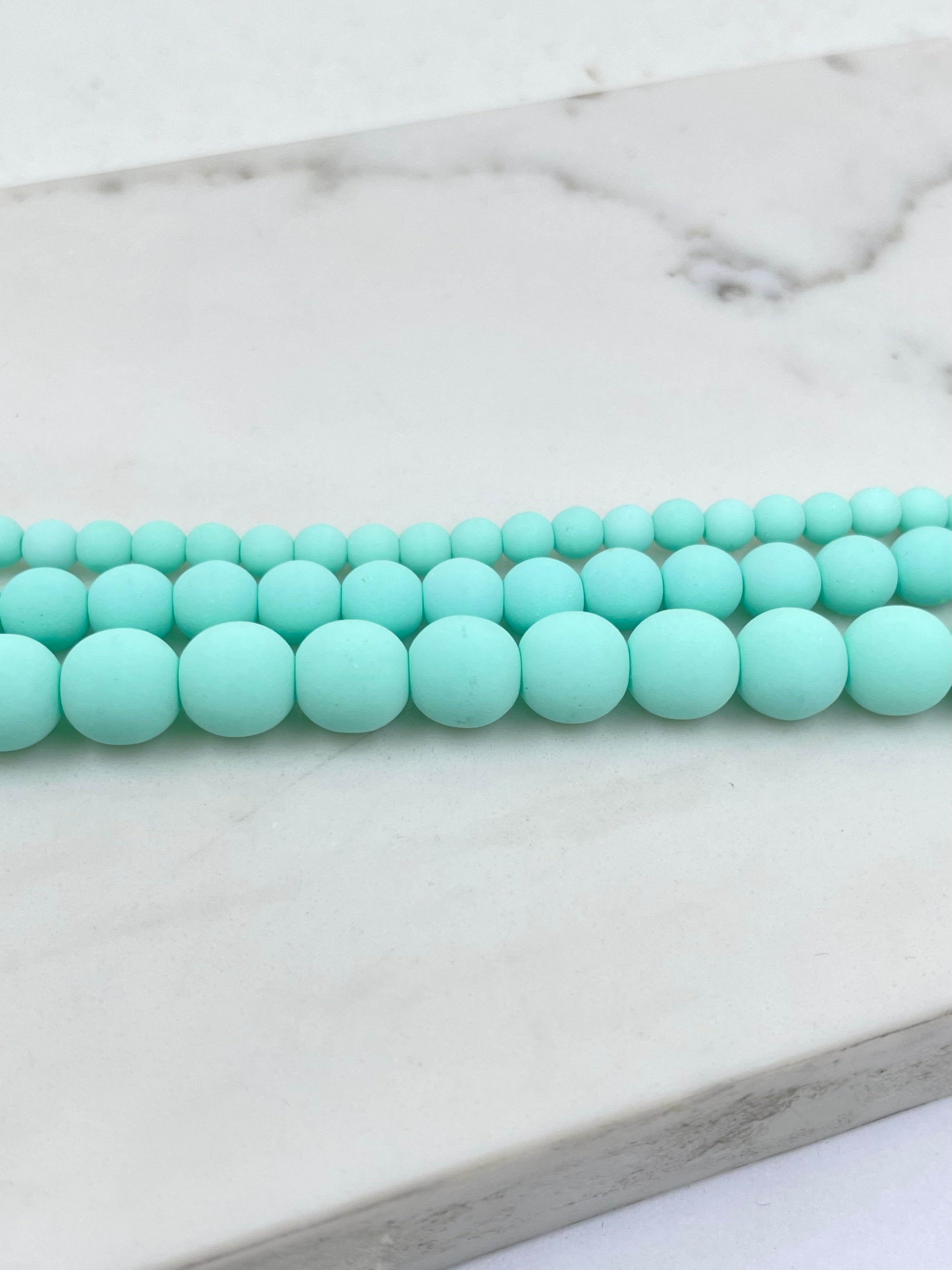 A close-up of seafoam blue matte round beads arranged in two rows on a white marble surface.
