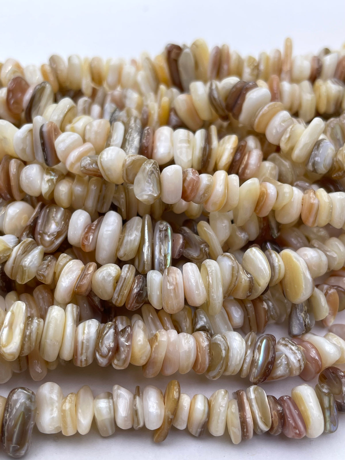 A close-up of multiple strands of small irregular shell beads in natural beige, cream, brown, and gray tones on a plain white background.