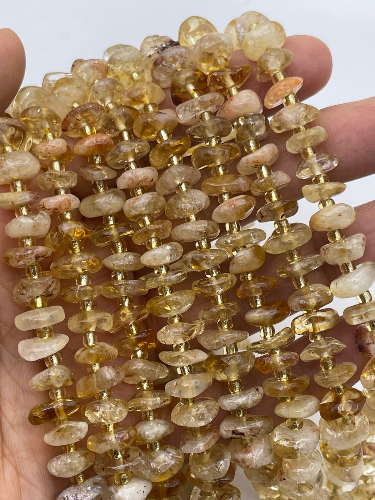 A close-up of a hand holding multiple strands of polished chunky citrine gemstone beads in warm amber, yellow, and cream tones with small gold spacer rings.