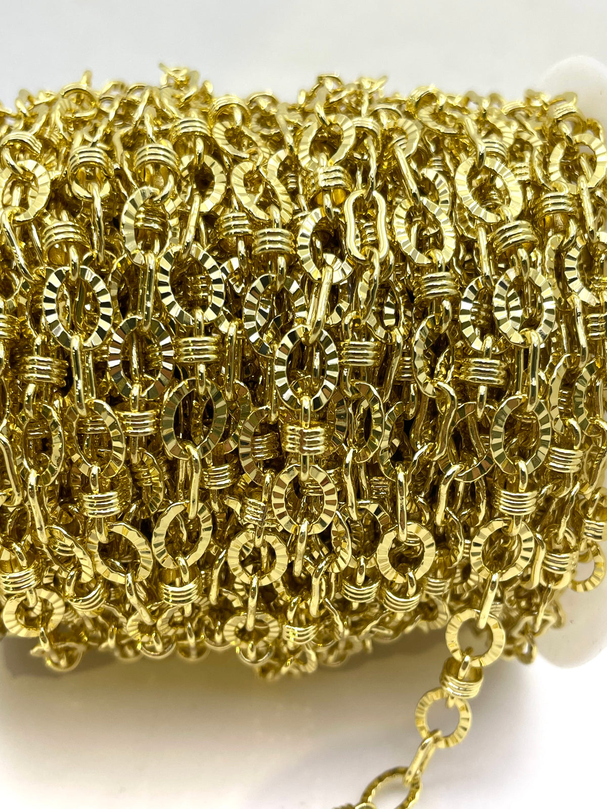 A close-up of a spool of gold-colored metal chain links, showing the detailed texture and reflective finish of the links on a white background.