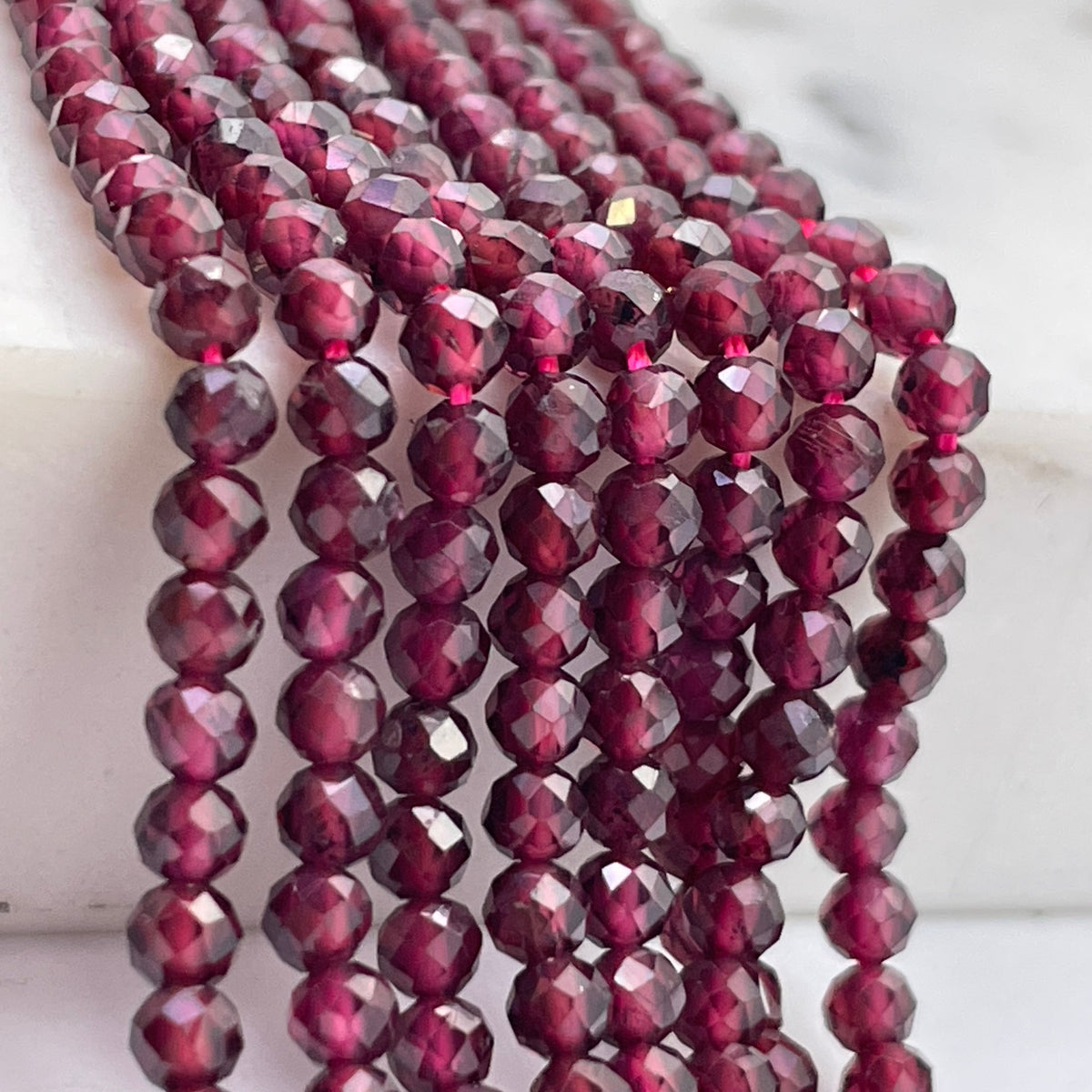 Faceted round garnet beads in a deep burgundy red color, arranged in multiple strands on a white marble surface.