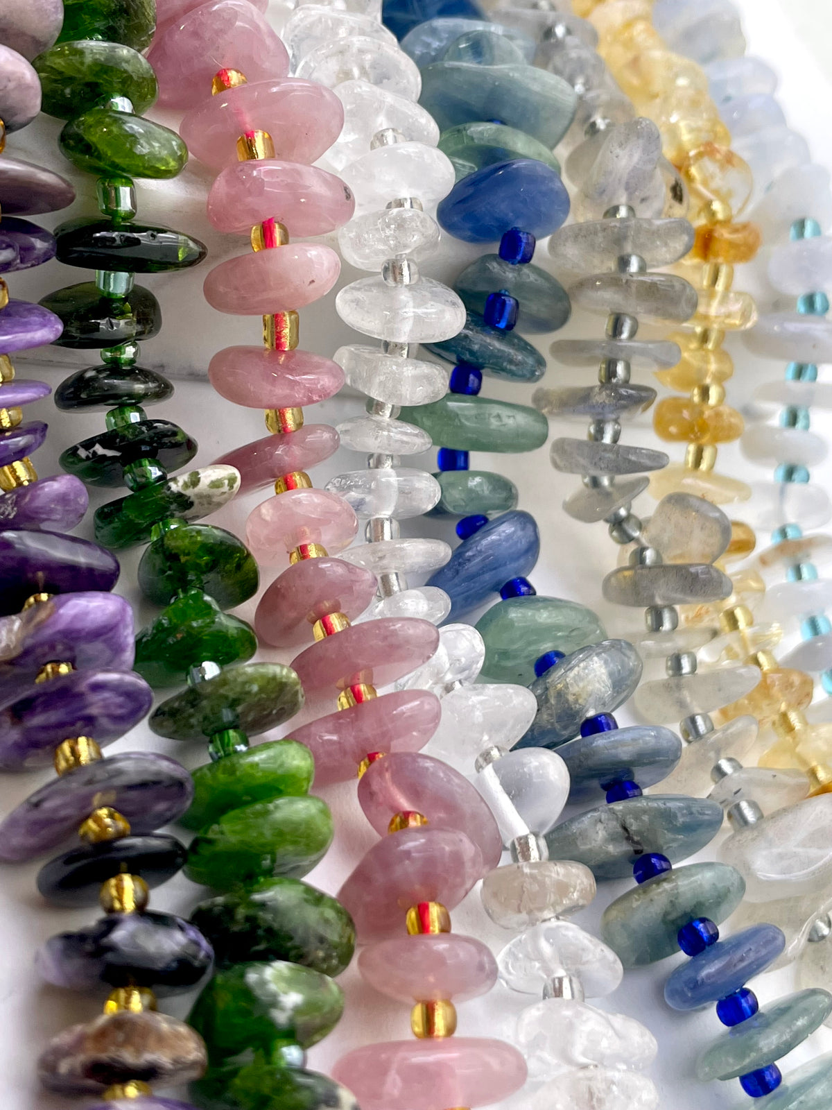 Close-up of multiple strands of polished stone beads strung on silver cords, arranged diagonally. The beads include pink, green, purple, blue, gray, clear, and yellow tones, with small metallic spacer accents.
