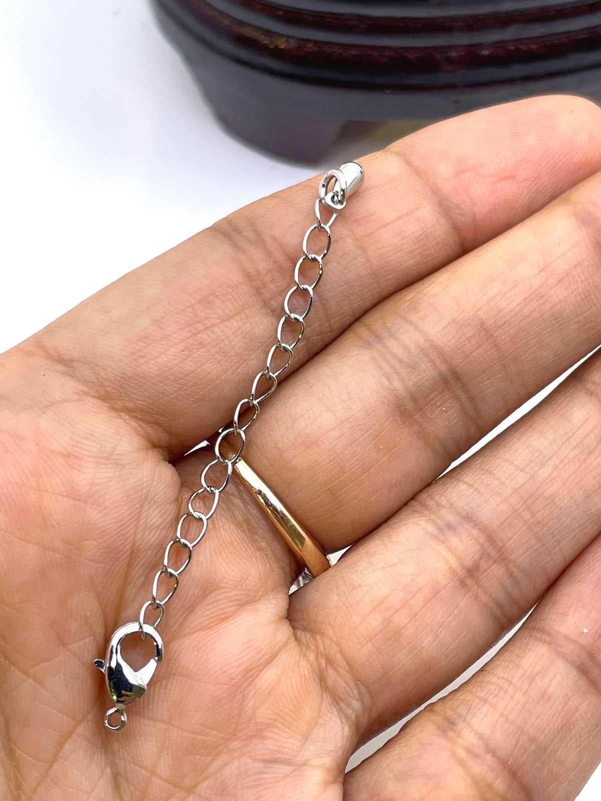A close-up of a hand holding a silver lobster clasp chain extender with a gold ring accent, showing the clasp detail against a white background.