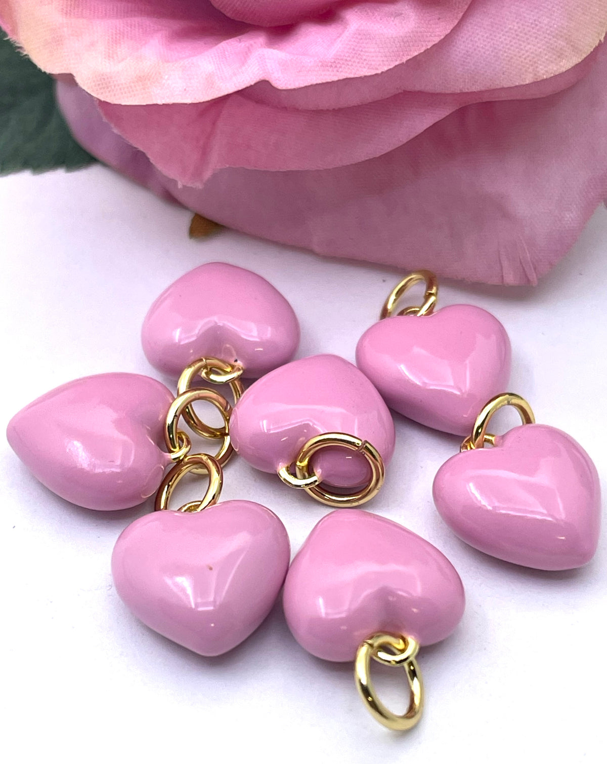 Six glossy pink heart-shaped beads with gold metal loops, arranged on a white surface with a soft pink rose in the background.