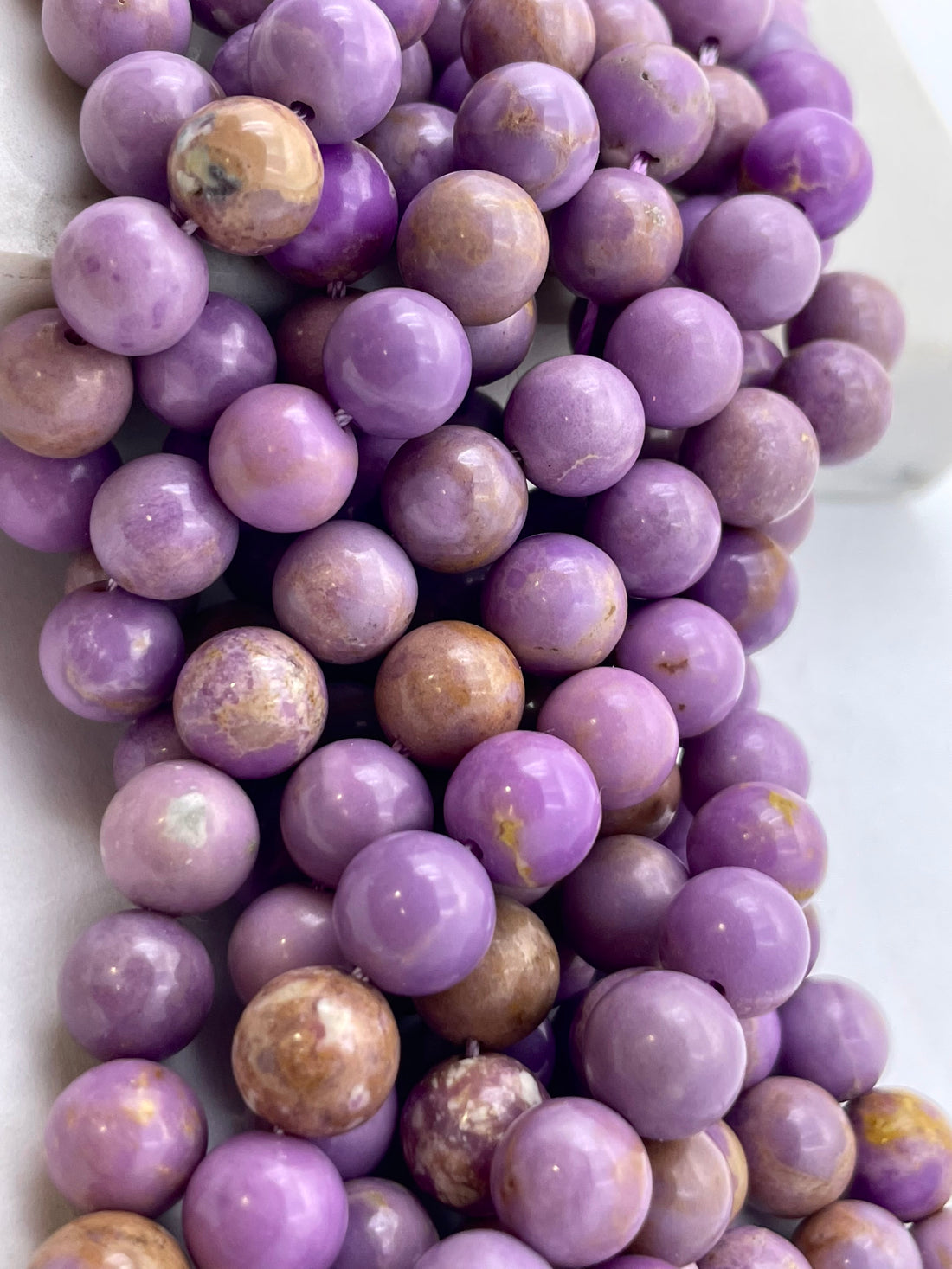 A close-up of a strand of polished  phosphosiderite lavender purple beads with natural brown and cream marbling, arranged on a light surface.