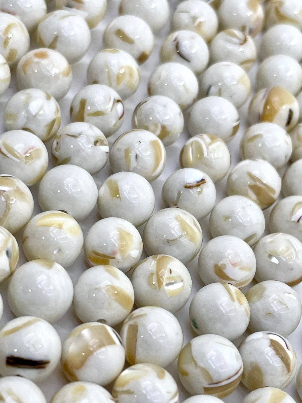 A close-up of many glossy round mop beads in an off-white marbled pattern with tan and brown swirls, filling the frame.