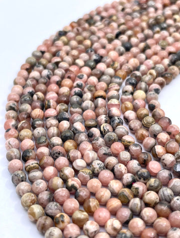 Curved strands of faceted round rhodochrosite beads made from pinkish stone with natural marbling, speckling, and dark inclusions, photographed on a white background.