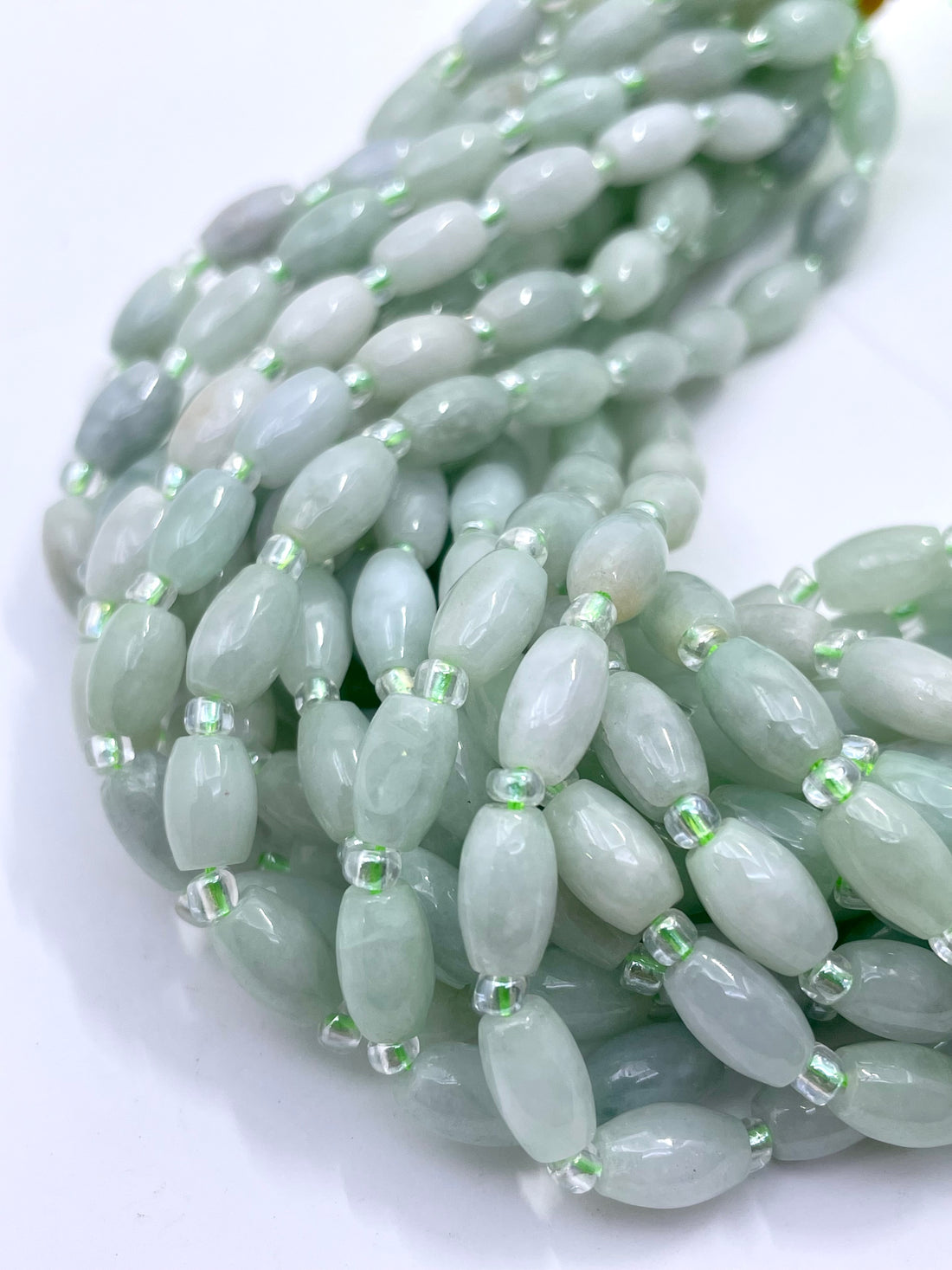 A hand holding multiple strands of  light green burma jade beads with small metallic spacer rings, shown against a plain white background.