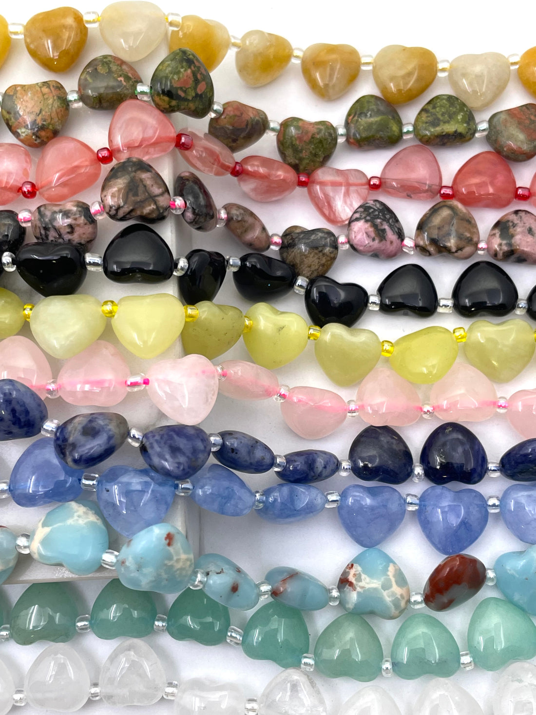 Multiple strands of polished gemstone heart-shaped gemstone beads arranged vertically, showing a variety of colors and stone types including pink, blue, green, yellow, black, and white.