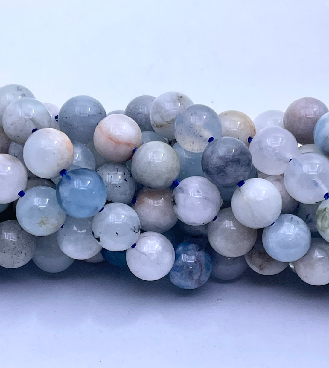 A close-up of polished round aquamarine gemstone beads in soft pastel shades of pale blue, white, gray, and faint peach, arranged on a textured gray surface.