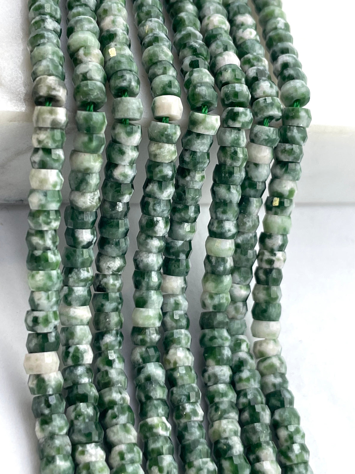 Multiple strands of faceted green spot jasper beads in green and white marbled stone, arranged vertically on a white background.